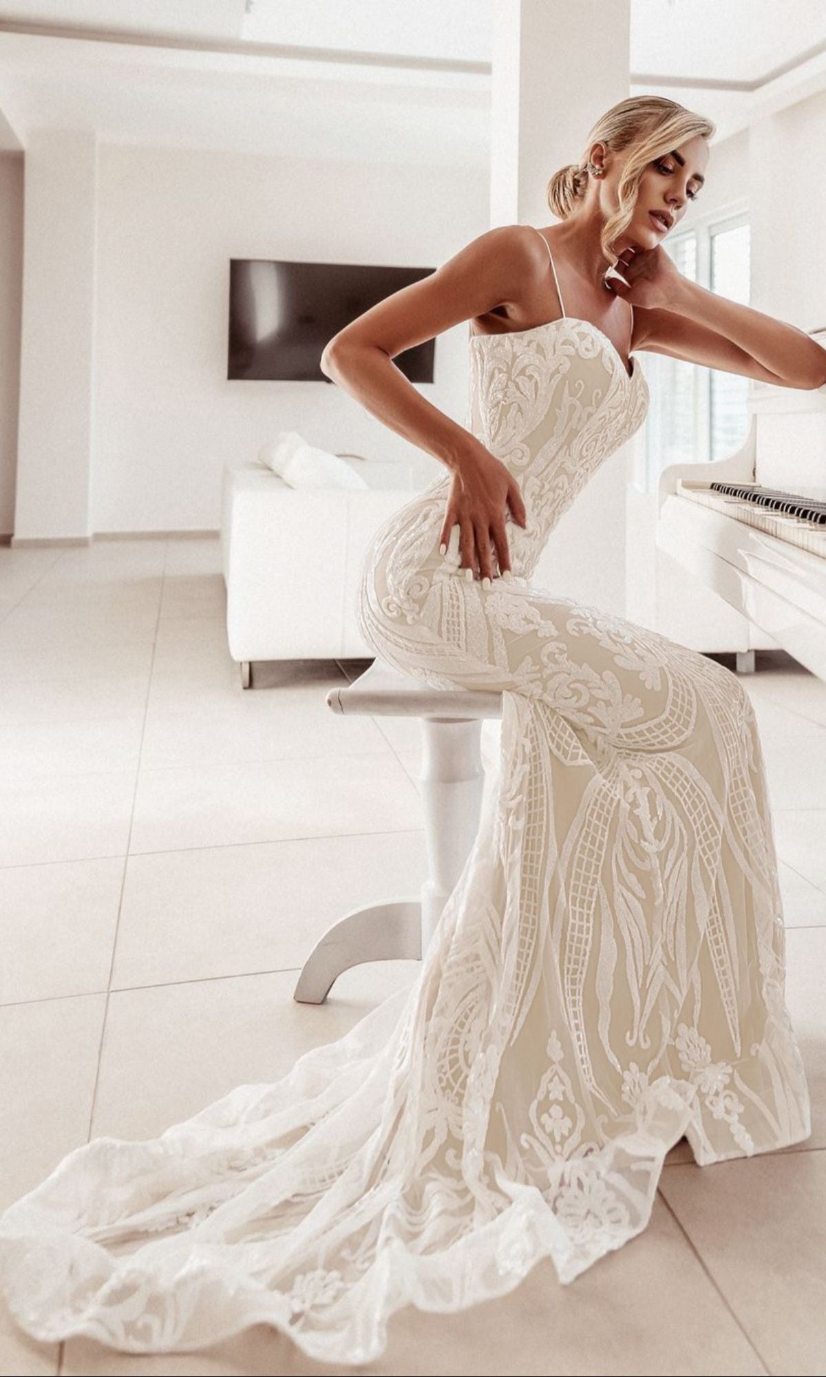 A woman wearing the White Lace Bodycon Bridal Gown: Tina Holly BA999 sits on a stool beside a white piano in a modern, minimalist living room.